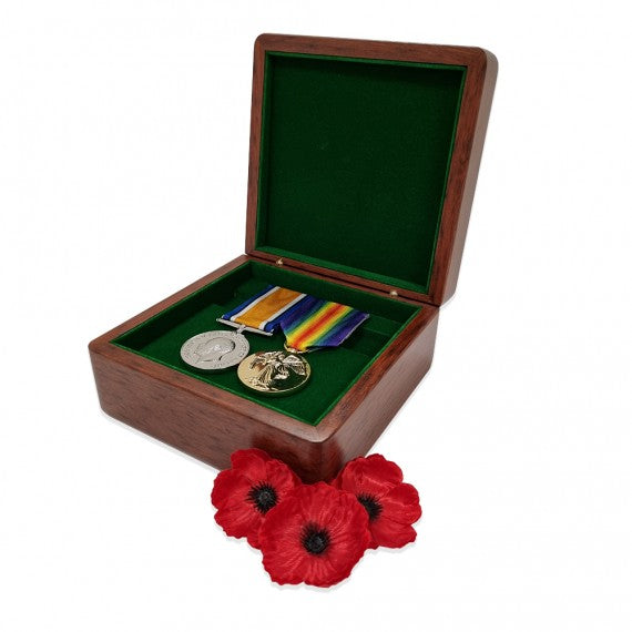 A wooden medal box with a green interior, featuring a silver and gold military medal displayed on a removable tray, accompanied by two poppy flowers outside the box.